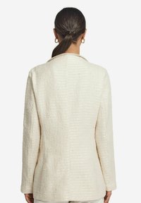 Ivoryfärgad kavaj med en subtil lyster, med slagkrage och långa ärmar, sedd bakifrån mot en vit bakgrund.