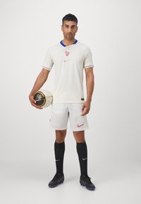 Maglia da calcio bianca con colletto blu, caratterizzata da un logo rosso e strisce. Abbinata a pantaloni corti bianchi, calze nere e un pallone da calcio nero e oro.