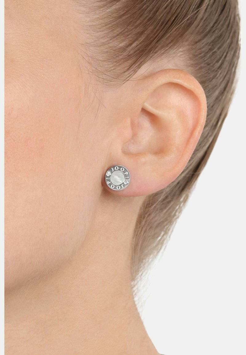 Silverfärgade studs i rund form, med en texturerad mitt och inristat varumärke, prydda med små klara stenar.