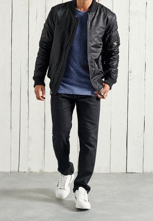 Veste aviateur noire au fini lisse, avec des poignets en maille et des poches zippées ; associée à un t-shirt bleu marine, un jean noir et des baskets blanches.