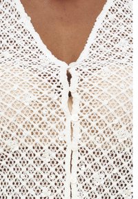 Gros plan sur une peau foncée portant un haut blanc en crochet avec de petits détails floraux et paillettes, partiellement boutonné au centre.