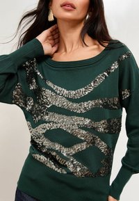 Zelený sveter s striebornými a čiernymi flitrovými vlnovými vzormi, so širokým výstrihom a rebrovanými detailmi na ramenách a manžetách.