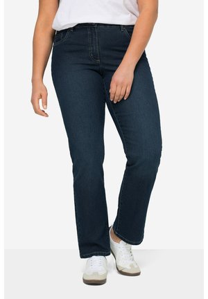 Persoon die donkere blauwe spijkerbroek en witte sneakers draagt, staande tegen een effen achtergrond.