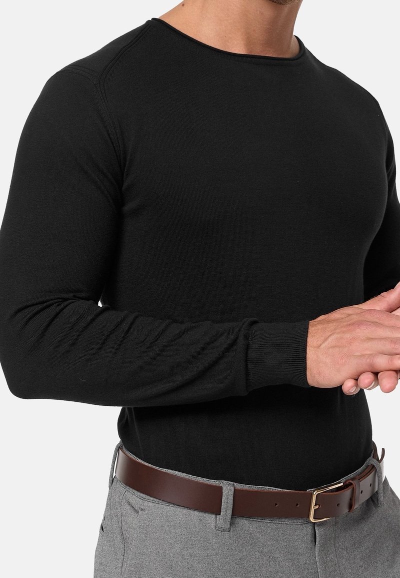 Homme portant un pull noir à manches longues, un pantalon gris et une ceinture en cuir marron, les mains jointes devant sur fond blanc.