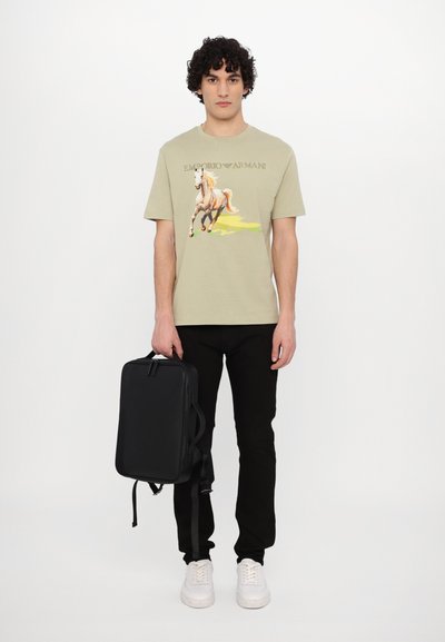 T-shirt kaki arborant un graphique de cheval avec le texte "Emporio Armani", accompagné d'un pantalon noir et d'un sac à dos noir rectangulaire. Des baskets blanches aux pieds.