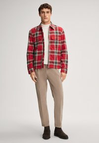 Młody mężczyzna w czerwono-szarej kurtce w kratę, białym prążkowanym golfie, beżowych spodniach i ciemnobrązowych butach, stojący na jednolitym tle.