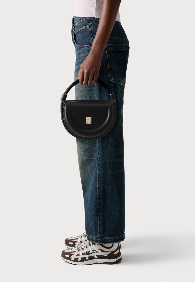 Sac à main en cuir noir avec une forme incurvée et une anse arrondie, présentant une petite plaque logo dorée. Porté avec un jean en denim bleu et des baskets.