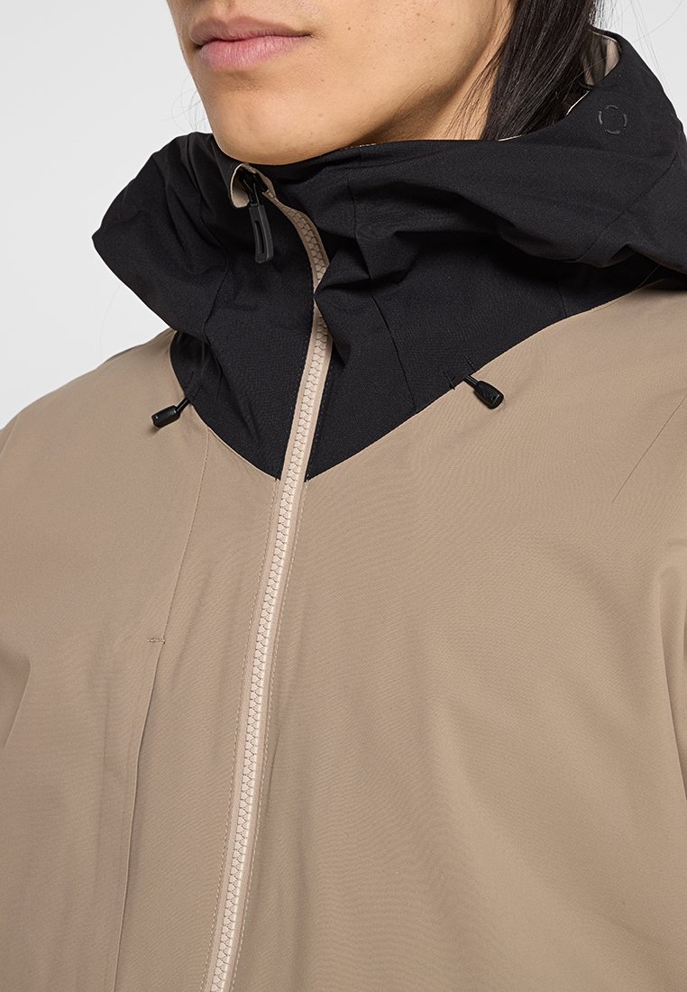 Nahaufnahme einer Person, die eine beigefarbene Jacke mit hohem, schwarzem Kragen und teilweise geschlossenem Reißverschluss trägt, wobei die glatte Textur des Stoffes zu sehen ist.