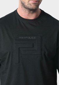 Camiseta negra con un diseño de logo en relieve y patrón grabado, fabricada en tela suave, con cuello redondo y mangas cortas.