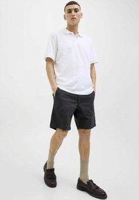Maglietta polo bianca a maniche corte, abbinata a pantaloni corti neri. Mocassini marroni e calzettoni beige completano l'outfit. Sfondo neutro.