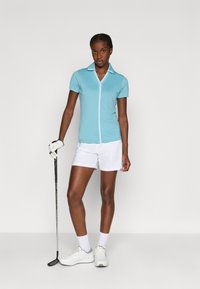 Svetle modré polo tričko se zipem vpředu a bílými detaily, spárované s bílými šortkami. Model drží golfovou hůl a má na sobě bílé boty se šedými akcenty.