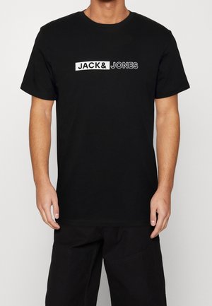 Homme portant un t-shirt noir à manches courtes avec l'inscription « JACK & JONES » imprimée en blanc et en contour sur la poitrine.
