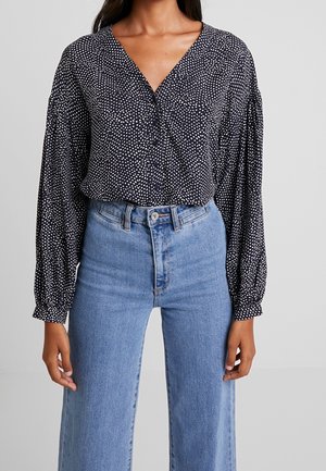 Frau trägt eine schwarze langärmelige Bluse mit weißen Punkten, in hoch taillierte hellblaue Jeans gesteckt.