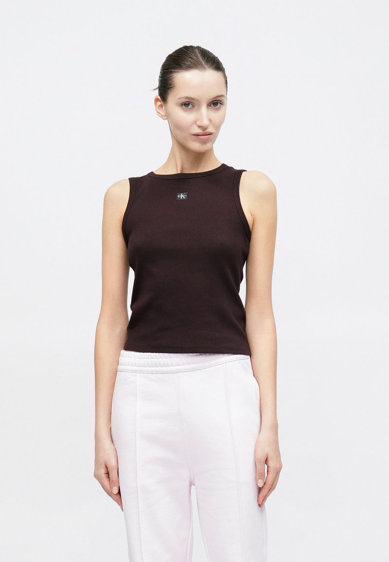 Σκούρο καφέ αμάνικο τοπ με ριμπ ύφανση, στρογγυλή λαιμόκοψη και μικρή λογότυπο λεπτομέρεια στο κέντρο μπροστά, συνδυασμένο με ανοιχτόροζ παντελόνι.