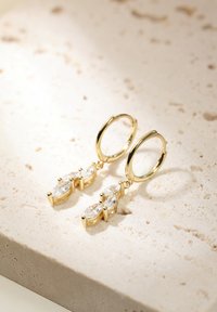 Goldene Creolenohrringe mit drei marquiseförmigen, klaren Steinen, die in einem goldenen Rahmen gefasst sind, und kleinen sphärischen Akzenten. Das Design ist modern und elegant.