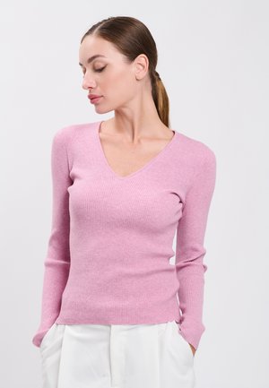 Frau mit braunen Haaren im Pferdeschwanz, die einen rosa gerippten V-Ausschnitt-Pullover und weiße Hose trägt, steht vor einem schlichten hellen Hintergrund.