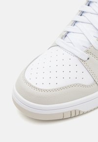 Baskets gris clair avec une tige en cuir lisse, un bout perforé blanc, des lacets blancs et une semelle en caoutchouc avec des rainures horizontales.