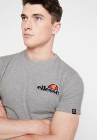 Gray t-shirt in cotton blend, featuring a round neckline. Embroidered "ellesse" logo in navy with a red and orange sun motif on the chest.