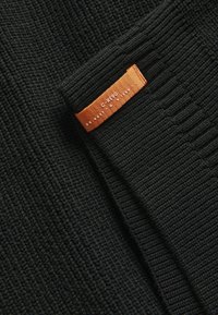 Tissu noir à côtes avec une surface texturée, arborant une étiquette orange avec du texte blanc. Le design global est simple et minimaliste.