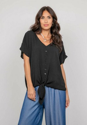 Femme aux longs cheveux bruns portant un haut noir ample boutonné et noué à la taille, avec un pantalon large bleu clair, sur un fond uni.