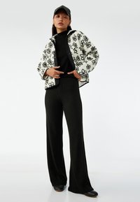 Fleecjasje met bloemen zwarte borduring, gecombineerd met een zwarte turtleneck en wijde zwarte broek. Zwarte pet en platte schoenen maken de look compleet.