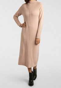 Robe tricotée rose clair avec un col montant côtelé et des manches longues, présentant une silhouette droite et des fentes latérales à l'ourlet. Bottines noires.