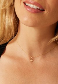 Collier en or avec un petit pendentif en forme de lettre 'M' texturée, posé sur une peau nue, avec des reflets et des ombres subtils. Design de chaîne simple.