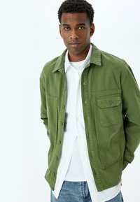 Mies, joka on pukeutunut vihreään napilliseen takkiin valkoisen paidan ja sinisten farkkujen päällä, seisoo valkoista taustaa vasten, kädet takin taskuissa.