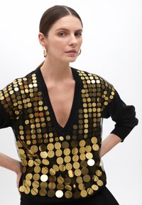 Black sweater featuring a deep V-neck, adorned with large gold sequins in a scale-like pattern. Long sleeves with ribbed cuffs.