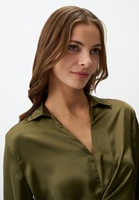 Blouse en satin vert olive avec un col, des manches longues et un détail drapé sur le devant. Texture lisse et éclat subtil.