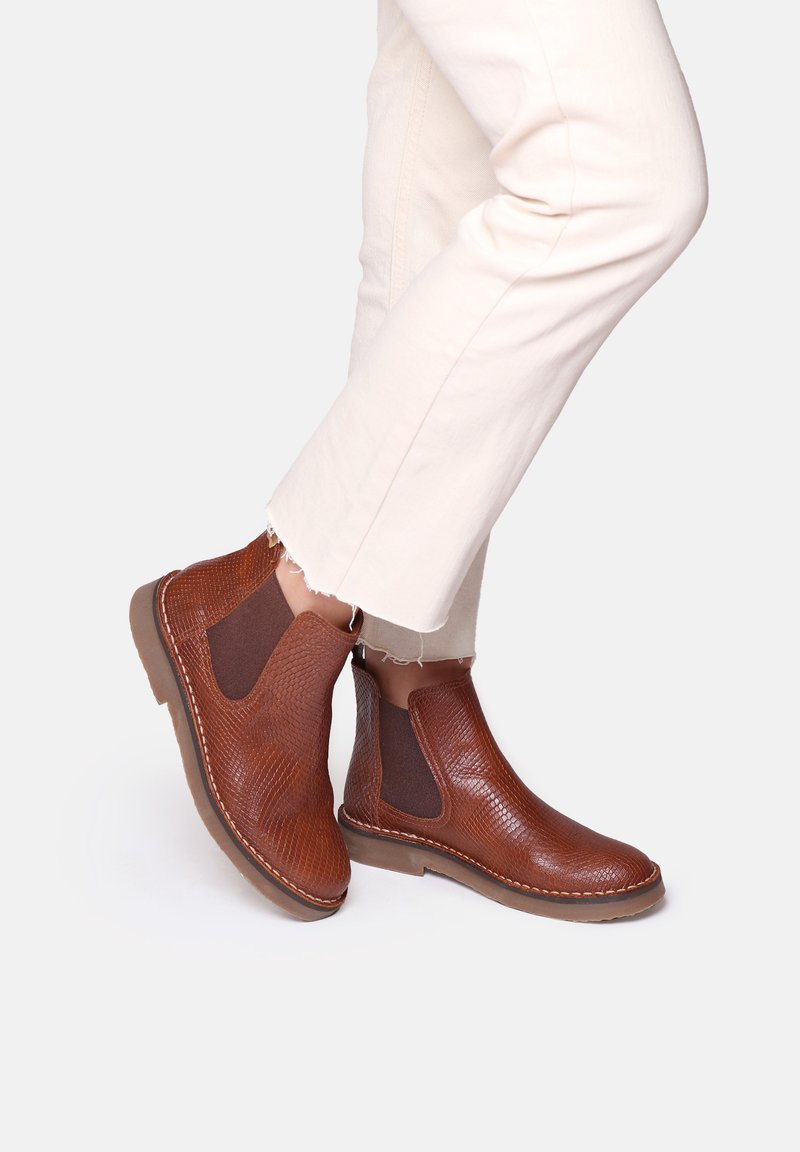 Botas Chelsea marrones hechas de cuero texturizado, con paneles laterales elásticos y suelas de goma, combinadas con pantalones de color crema.
