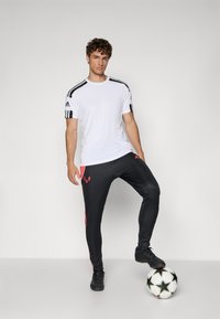 White athletic t-shirt with black stripes on shoulders, paired with black pants featuring red accents, standing on a black and white soccer ball.