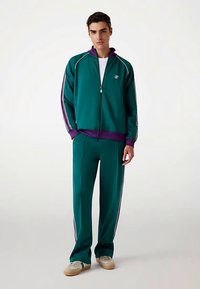 Grön sportoverall med lila krage och ränder på sidorna, zip-up jacka och matchande byxor med raka ben.