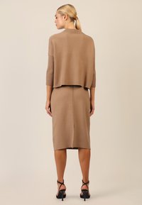 Strick-Zweiteiler in Hellbraun mit einem kurzen Oberteil mit gerippten Ärmeln und einem taillierten Rock mit einem Rückenschlitz. Schwarze Riemchen-Heels.