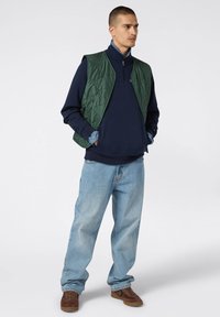 Gilet imbottito verde sopra un felpa blu navy, abbinato a jeans azzurri e sneakers marroni. Look casual a strati con colori contrastanti.