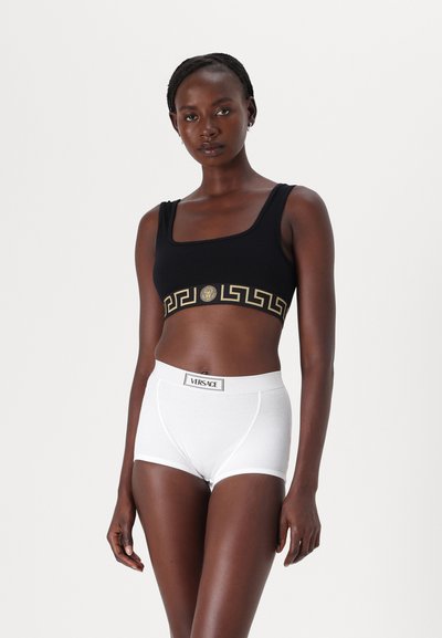 Mustat lyhythihaiset crop-topit, joissa on kultainen kreikkalainen avainkuvio ja logo, yhdistetty valkoisiin ribbimuotoisiin shortseihin, joissa on brändätty vyötärö.