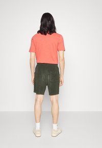 T-shirt rouge à manches courtes et short vert olive avec poche arrière. Personne portant des baskets beiges et des chaussettes blanches, vue de dos.