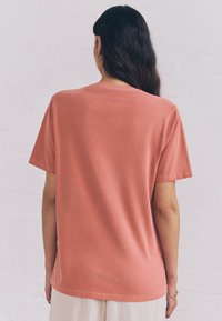 Woman with long dark hair wears coral short-sleeve t-shirt, gold hoop earrings, and a bracelet, standing against a white brick wall.