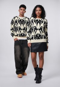 Strickpullover in Creme mit abstraktem schwarzen Muster; das Model kombiniert ihn mit einem schwarzen Lederrock und hohen schwarzen Stiefeln. Das männliche Model trägt ausgewaschene Jeans.