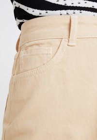 Pantalones de algodón color beige claro con un acabado texturizado, que cuentan con un bolsillo frontal y una cintura con un pequeño lazo para el cinturón.