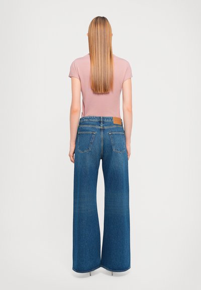 Jean en denim à jambe large de couleur bleu foncé avec un effet délavé, présentant une étiquette en cuir au dos de la taille et deux poches arrière.