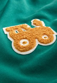 Un patch de tracteur orange texturé cousu sur un tissu vert avec des coutures visibles autour de la bordure blanche.