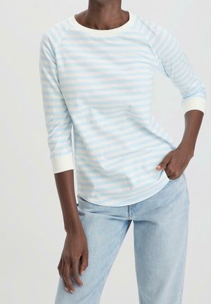 Person iført lyseblå og hvidstribet langærmet skjorte med hvide manchetter og lyseblå jeans, den ene hånd i lommen.