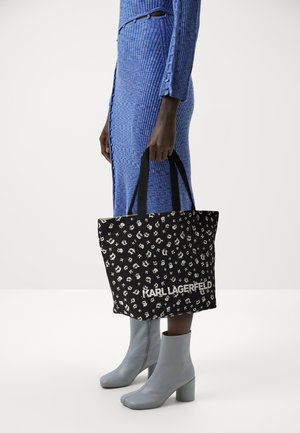 Svart tygväska med vitt mönster, med logotypen "KARL LAGERFELD", robusta handtag och rektangulär form. Modellen bär en blå ribbad klänning.