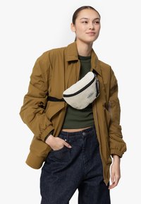 Marsupio bianco in corduroy con zip nera, indossato sopra una maglia verde oliva e un giubbotto in piuma marrone, abbinato a pantaloni in denim blu scuro.