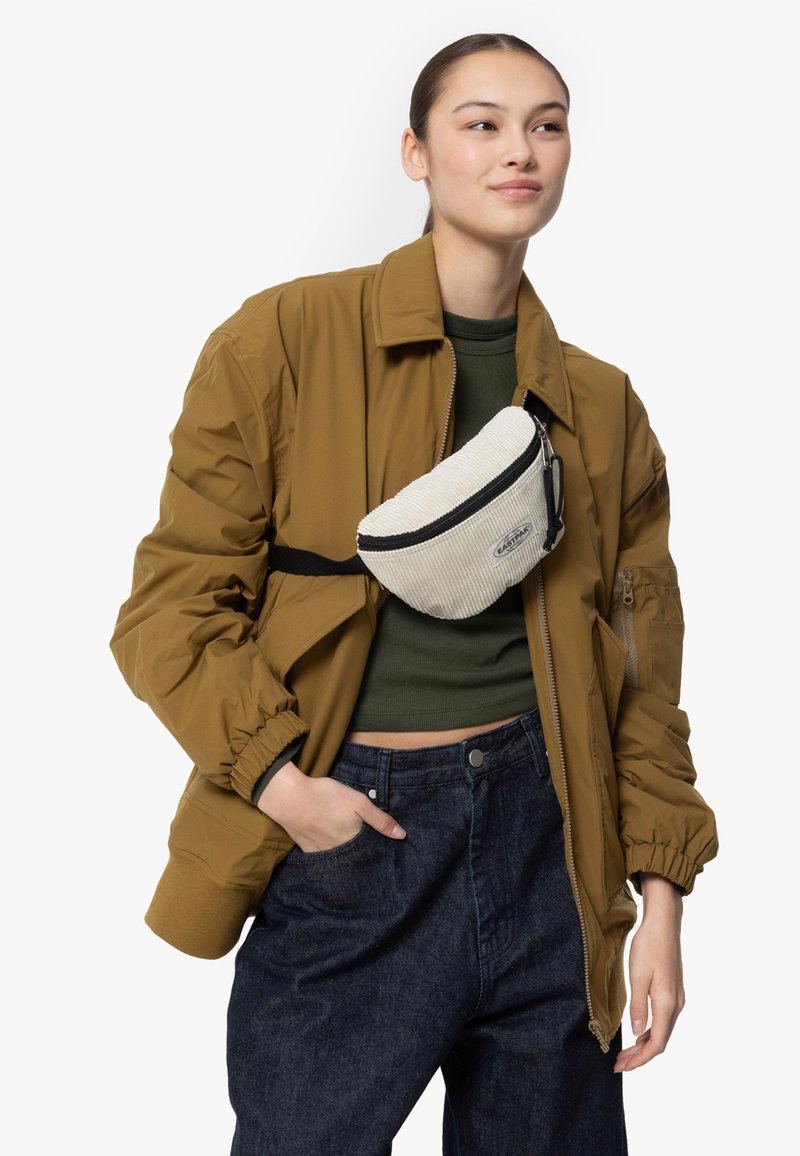 Marsupio bianco in corduroy con zip nera, indossato sopra una maglia verde oliva e un giubbotto in piuma marrone, abbinato a pantaloni in denim blu scuro.