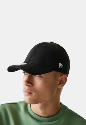 Uomo che indossa un berretto da baseball nero con un logo bianco sul lato, guarda a sinistra, vestito con una maglietta verde girocollo.