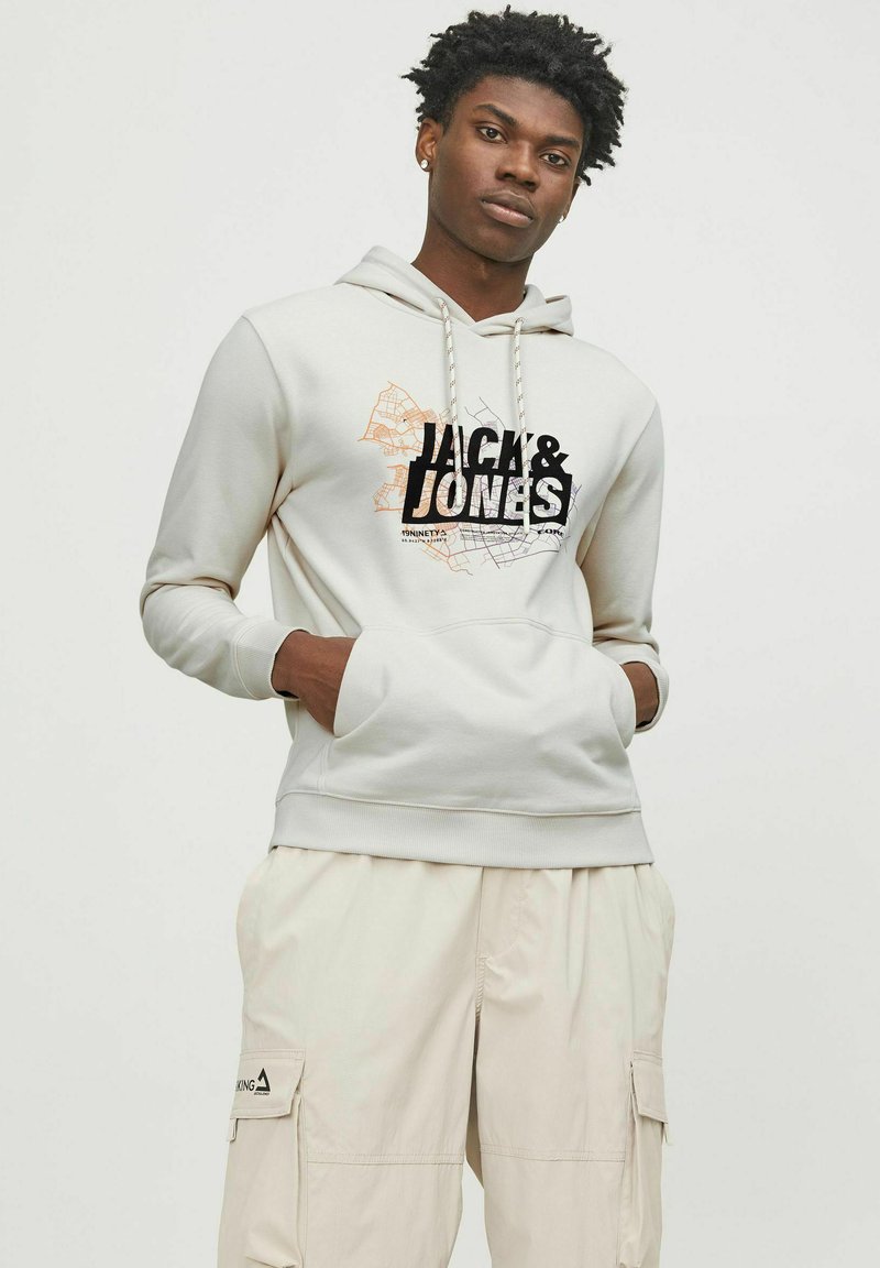 Lysegrå hættejakke med grafik foran, som viser "JACK & JONES" i sort og orange. Materiale: blødt, glat stof; justerbar snor i hætten.