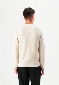 Mann med kort mørkt hår iført en teksturert off-white langermet genser og svarte bukser, vendt bort fra en hvit bakgrunn.