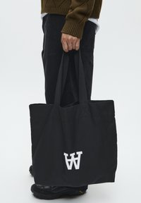 Black canvas tote bag with contrasting white logo, featuring sturdy handles and a minimalistic design, held in a person's hand.